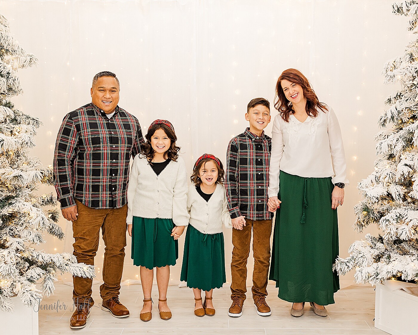 Family standing and holding hands by a light wall and Christmas trees for a Christmas studio family portrait session near Erie, PA.