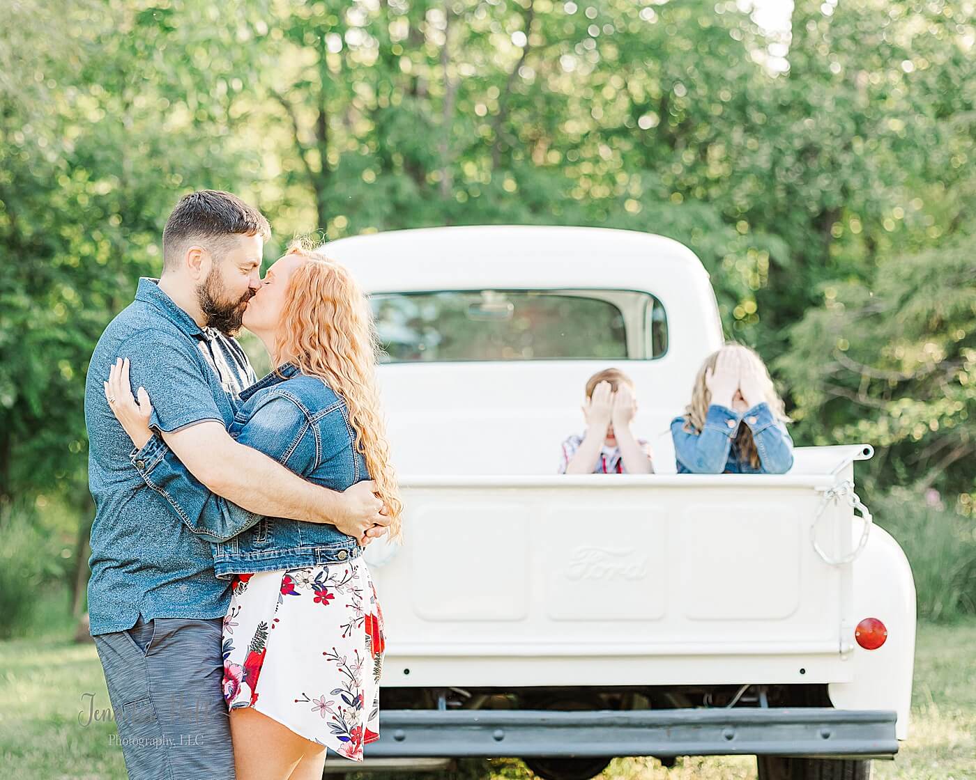 Mother and father kissing in front of a truck with their kids sitting in the truck and closing their eyes for summer family photos with a vintage truck near Erie, PA.