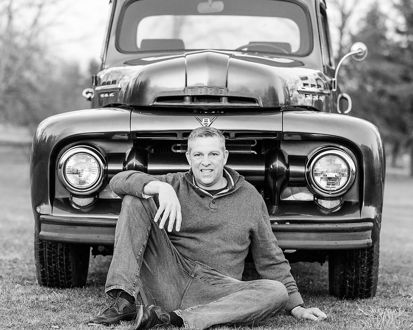 Father with a sweater sitting between the headlights of a truck in the grass outdoors.