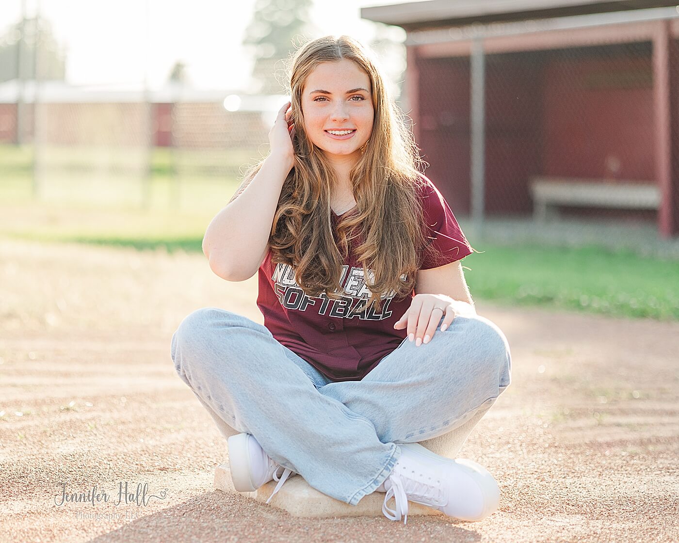 Girl with her jersey sitting on a softball base outdoors at a softball field for summer senior photos in North East, PA.