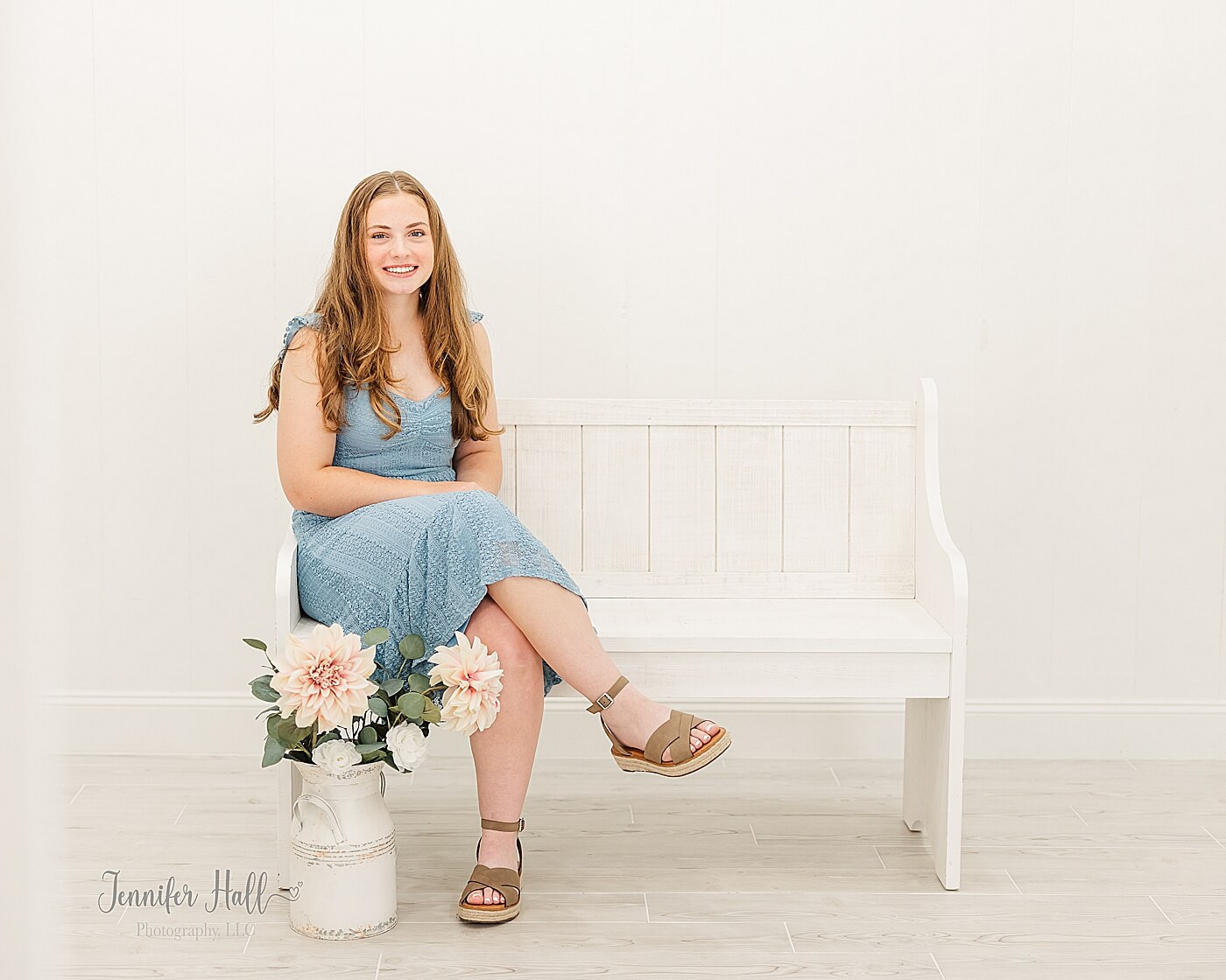 Girl in a blue dress sitting on a white wooden bench with flowers in a photo studio for summer senior photos in North East, PA.