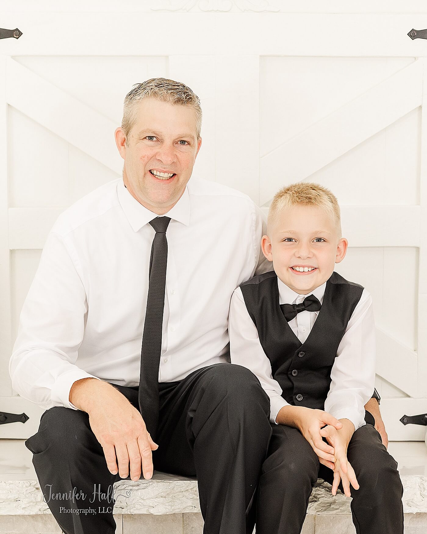 Father with a white solid, collared shirt with a black tie and black dress pants, sitting next to his son who is wearing a white solid, collared shirt with a black suit vest, a black bow tie, and black dress pants to show what to wear for him for family photos.