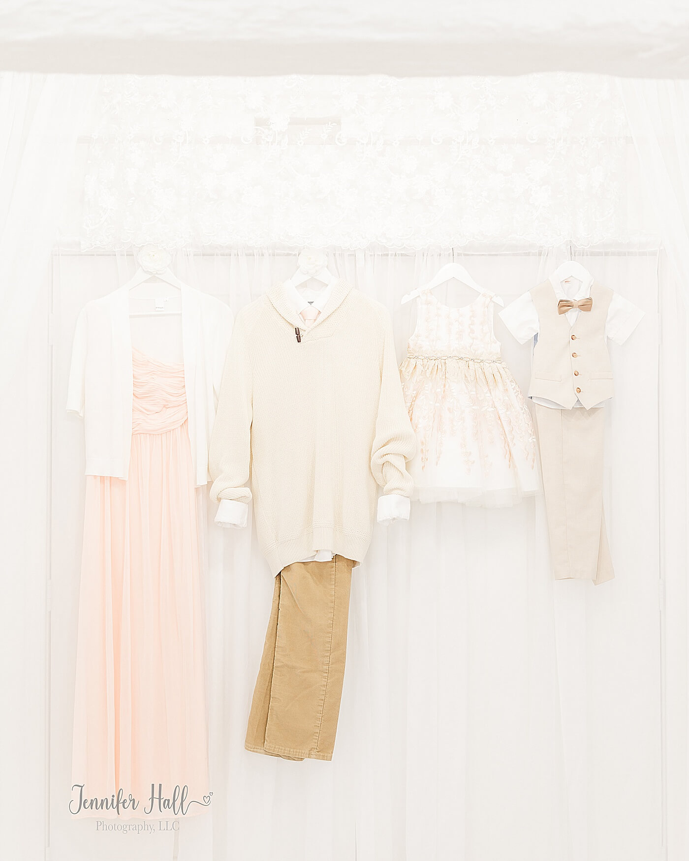 Vanilla blush dress with a white sweater, a beige sweater with tan corduroys, a little girl’s white dress, and a little boy’s beige suit vest and tan bow tie hanging up in a studio to show what to wear for family photos.