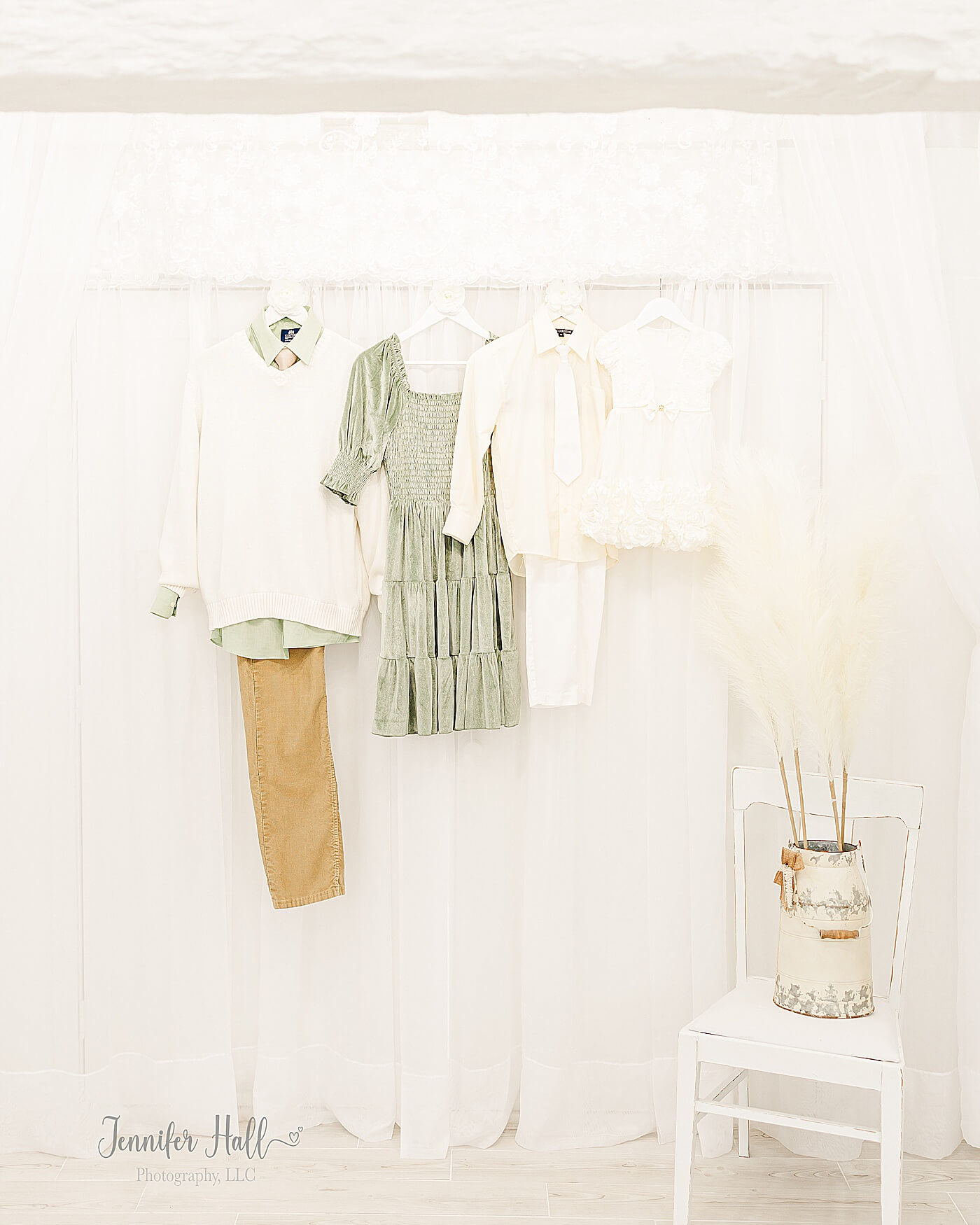 Dusty sage dress; a dusty sage dress shirt with a white sweater vest and a wafer tie; a cream little girl dress; a pale-yellow dress shirt with a white tie hanging on a white wall for a dusty sage and white fall family outfits.