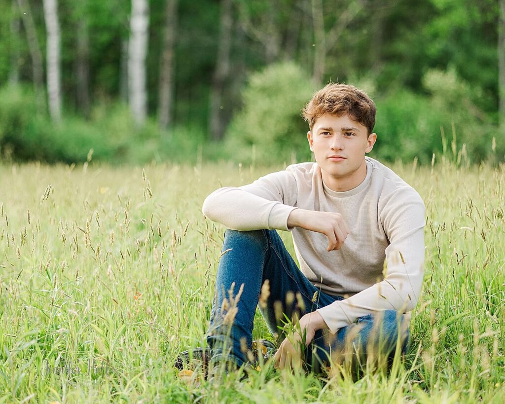 Senior guy with a cream shirt sitting in tall grass in the woods for summer senior pictures in Allegany, New York.