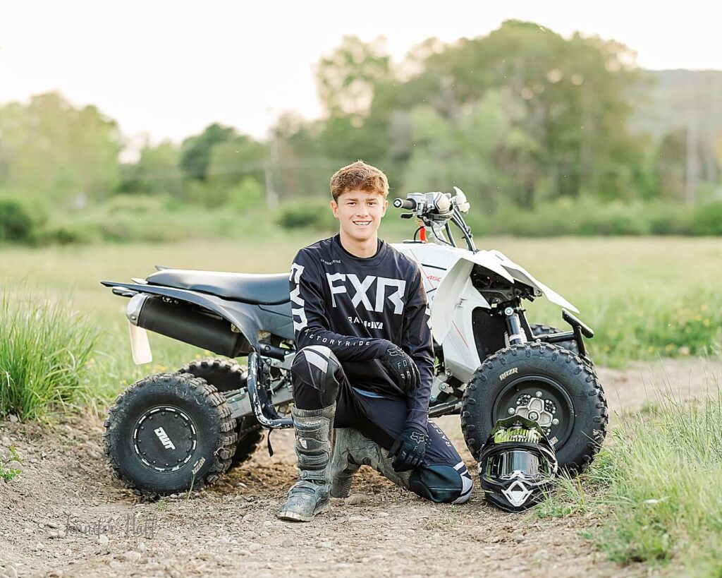 Senior guy kneeling by the side of his four-wheeler outdoors on a dirt track for summer senior pictures in Allegany, New York.