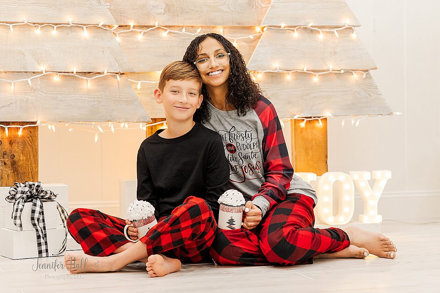 Brother and sister with hot cocoa mugs by wooden Christmas trees for indoor Christmas family photos.