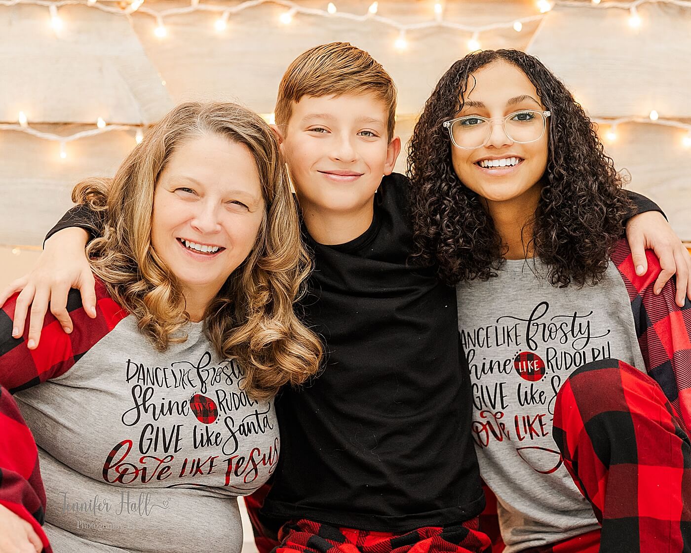 Family smiling with their arms around each other by wooden Christmas trees for Christmas family photos in-studio.