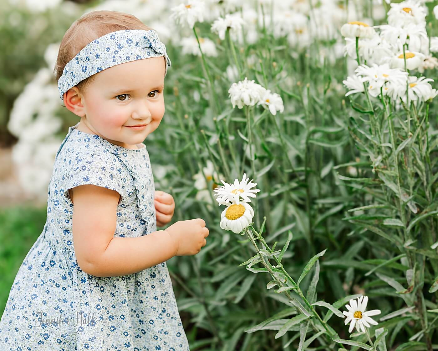 Little girl smiling by white daisies outdoors.