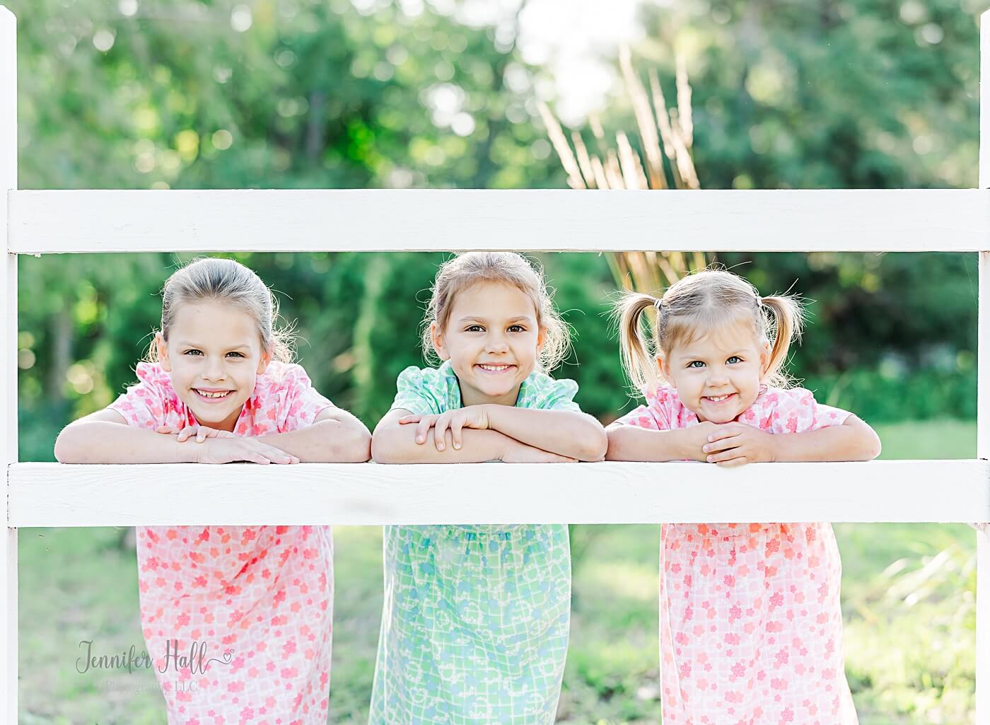 Girls with pink and green dresses standing by a white fence and smiling.