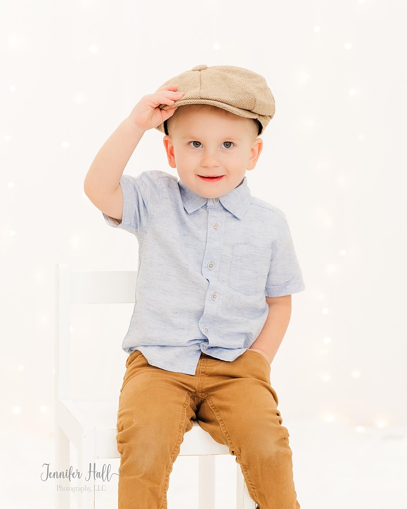 Little boy holding a beige newsboy cap and sitting in a white chair.