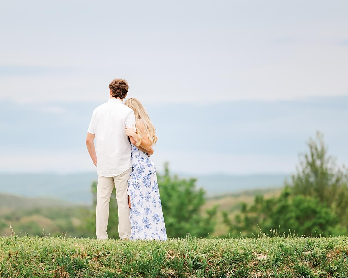 Man hugging his fiancée, facing a mountain view, for an engagement photo session.