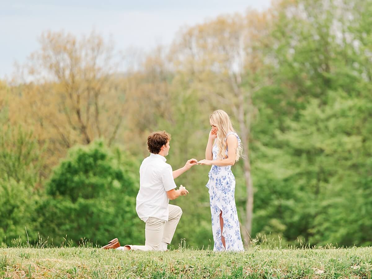 Man placing an engagement ring on a woman’s finger by a pond for engagement photos.